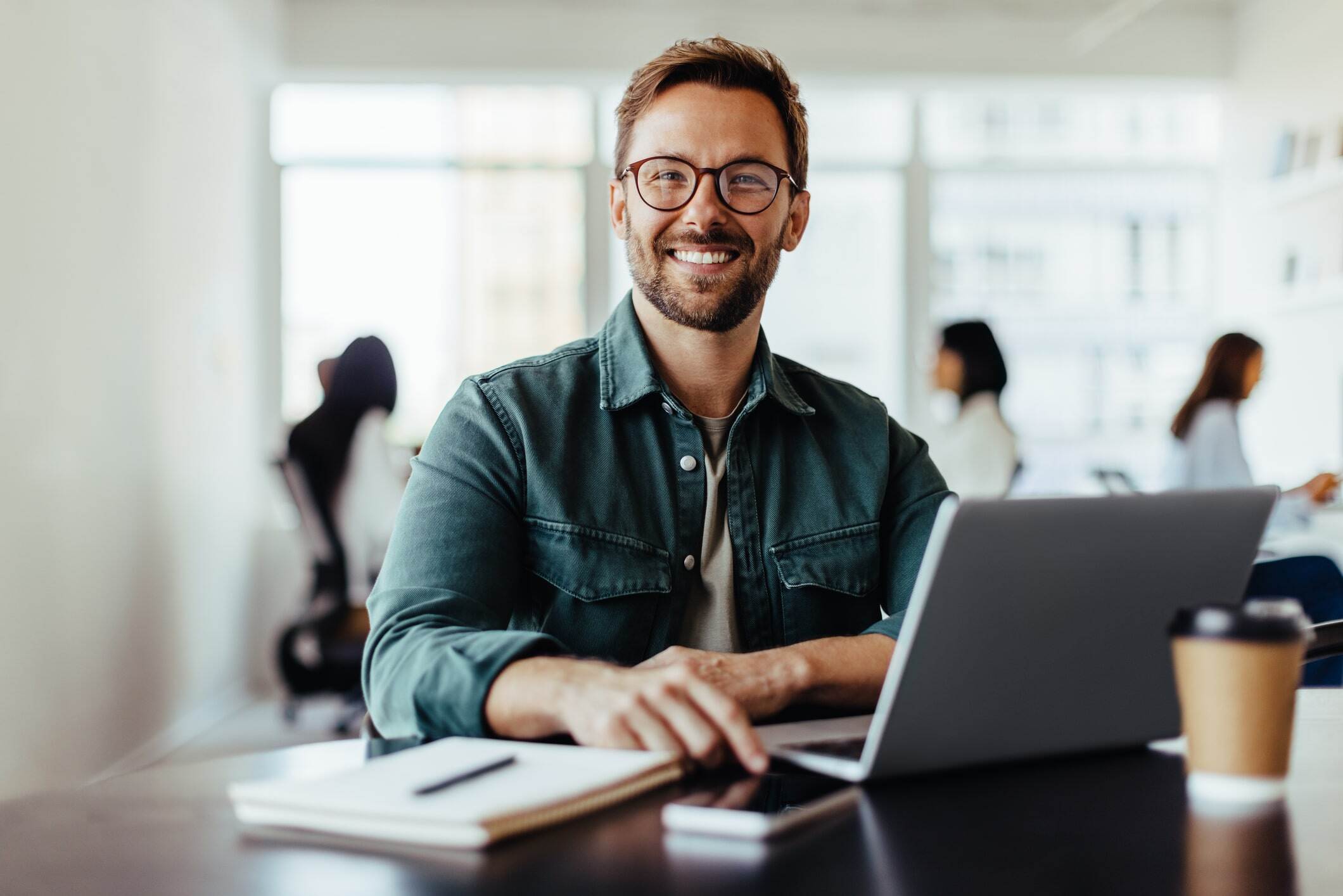 Smiling man with glasses sits at a desk in the office and uses a computer for time tracking.